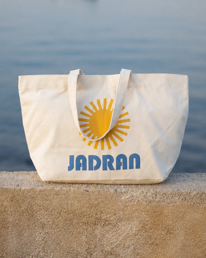 A cream canvas tote bag with a yellow sun graphic and the word 'JADRAN' in blue, resting on a stone wall with a blurred body of water and distant boat in the background.
