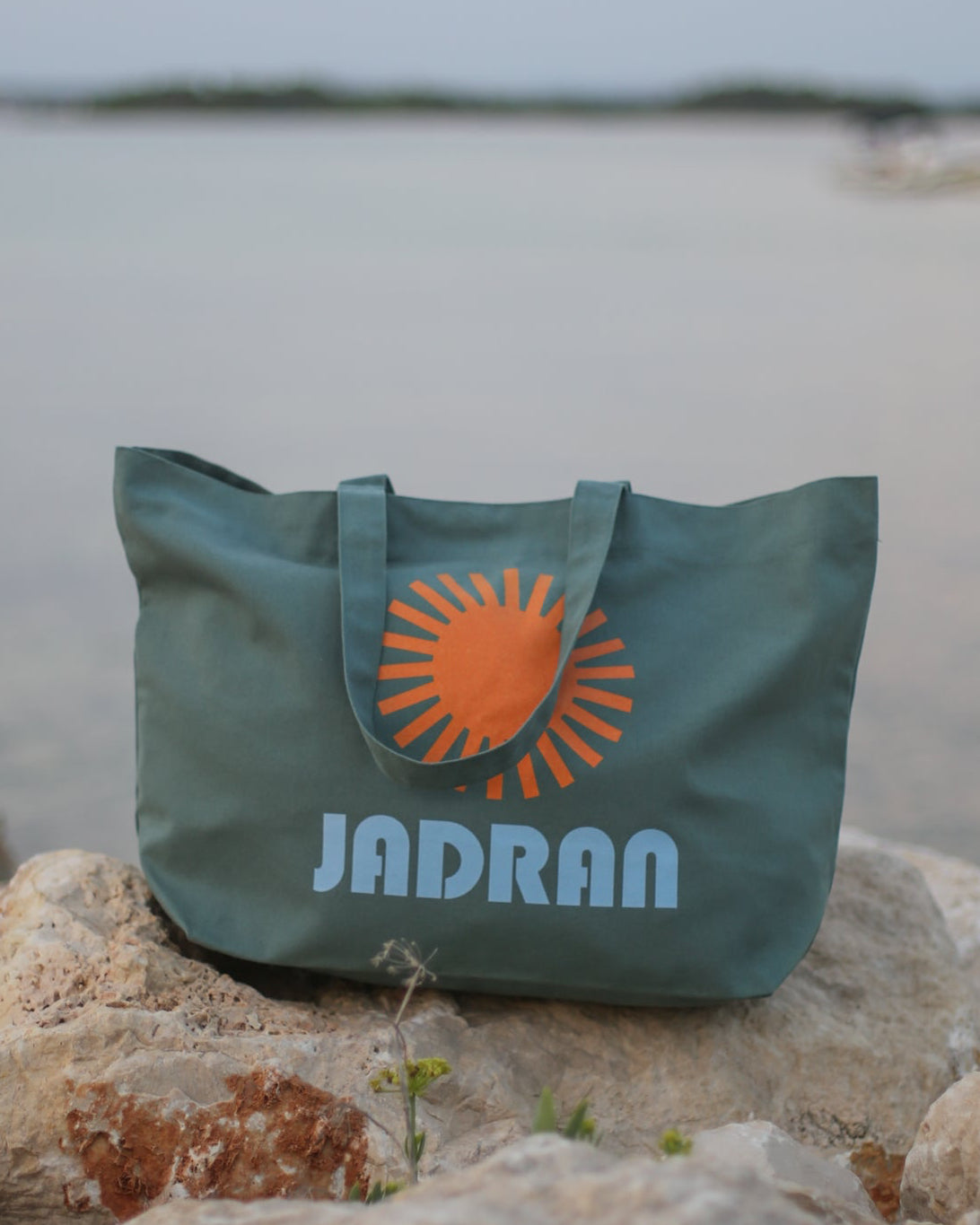 A green tote bag with an orange sun logo and the brand name 'JADRAN' printed in light blue, resting on rocks with a blurred lake and sky background.