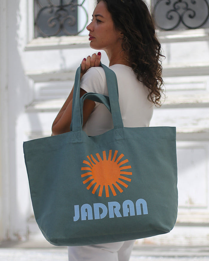 A woman in a white outfit carries a large slate blue tote bag with an orange sun graphic and the text 'JADRAN' in light blue, standing in front of a white building with decorative windows.