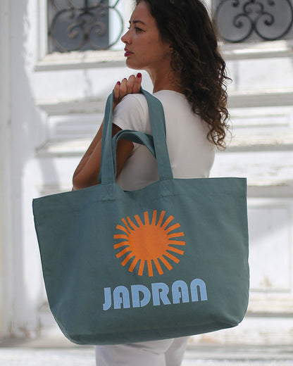 A woman in a white outfit carries a large slate blue tote bag with an orange sun graphic and the text 'JADRAN' in light blue, standing in front of a white building with decorative windows.