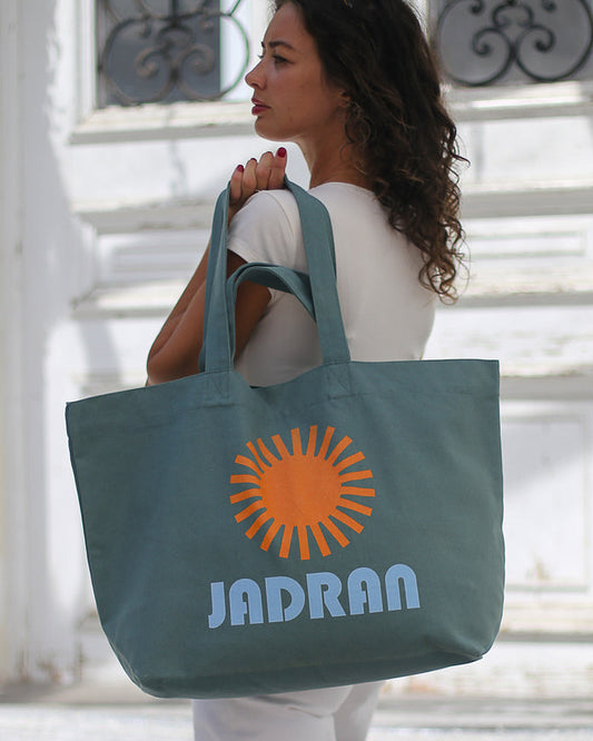 A woman in a white outfit carries a large slate blue tote bag with an orange sun graphic and the text 'JADRAN' in light blue, standing in front of a white building with decorative windows.