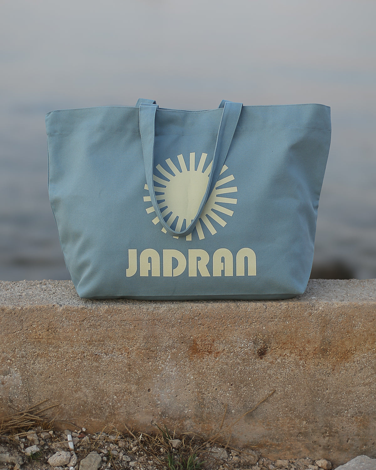 A light blue canvas tote bag with a cream-colored sun graphic and the brand name 'JADRAN' printed on the front, resting on a concrete ledge with a blurred outdoor background.