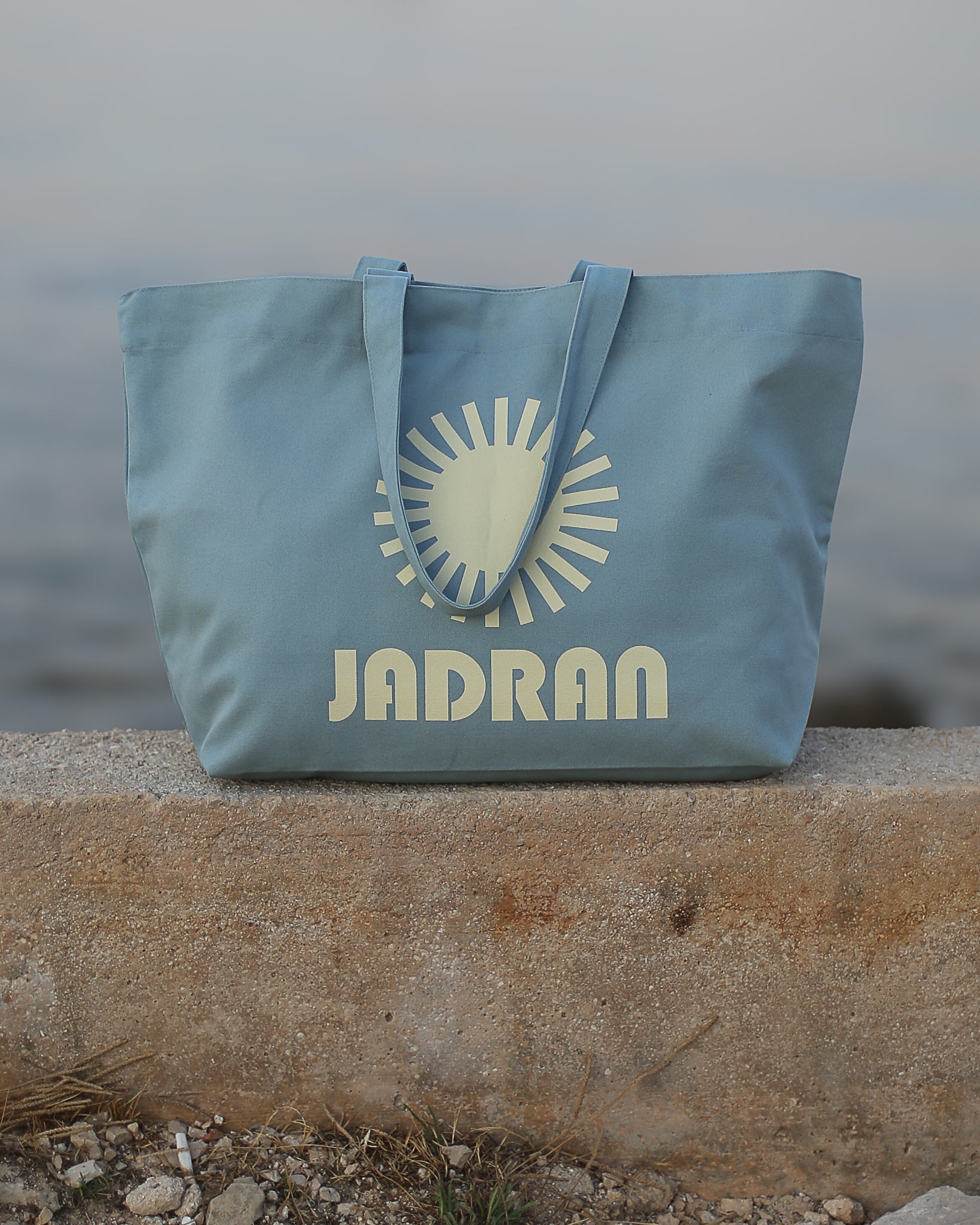 A light blue canvas tote bag with a cream-colored sun graphic and the brand name 'JADRAN' printed on the front, resting on a concrete ledge with a blurred outdoor background.