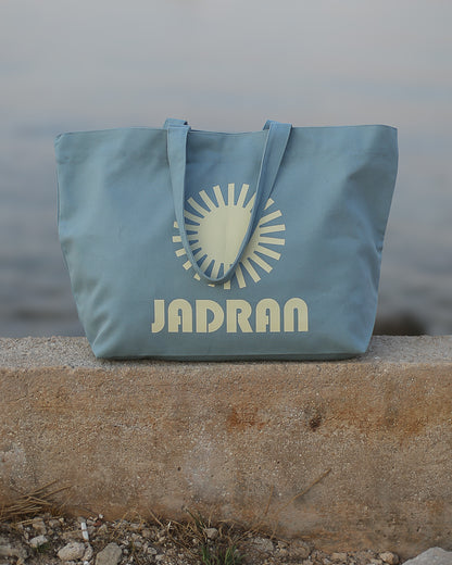 A light blue canvas tote bag with a cream-colored sun graphic and the brand name 'JADRAN' printed on the front, resting on a concrete ledge with a blurred outdoor background.